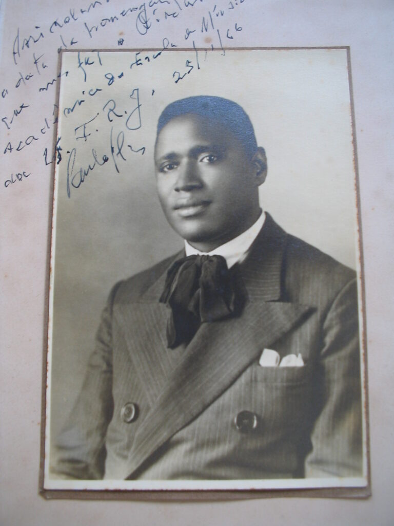 Homem negro com cabelo preto e crespo, vestido de paletó preto com blusa branca e gravata preta em formato de laço. A foto está em uma coloração sépia. Está em uma postura em que o corpo está levemente inclinado para  o lado direito e seu olhar está voltado para frente. 