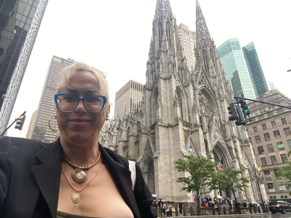 Imagem 1 - Mulher branca, cabelo liso e loiro preso para trás. Está usando um óculos em armação azul, e brincos de argolas dourados. Usa dois colares dourados com pingentes redondos  e blazer preto. Ao fundo há uma catedral em estilo gótico, um sinal de trânsito marcando a cor verde e prédios ao fundo. 