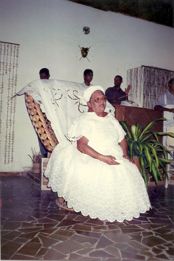 Imagem 2 - mulher branca, idosa, sentada em cadeira de palha coberta com pano branco bordado. Ela veste roupa de axé branca rendada  e turbante branco cobrindo o cabelo. está sentada com as mãos apoiadas em suas pernas. Usa óculos. Chão de revestido de cacos de azulejos marrons formando uma espécie de mosaico. em su lado esquerdo há uma planta e ao fundo quatro homens negros, um deles tocando atabaque.