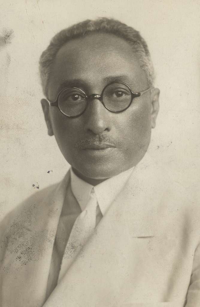 Imagem - Foto em preto e branco  de homem negro sério, com óculos redondo em armação preta, cabelo grisalho e curto e bigode grisalho. veste um terno, blusa e gravata brancos. Fundo da imagem branco.