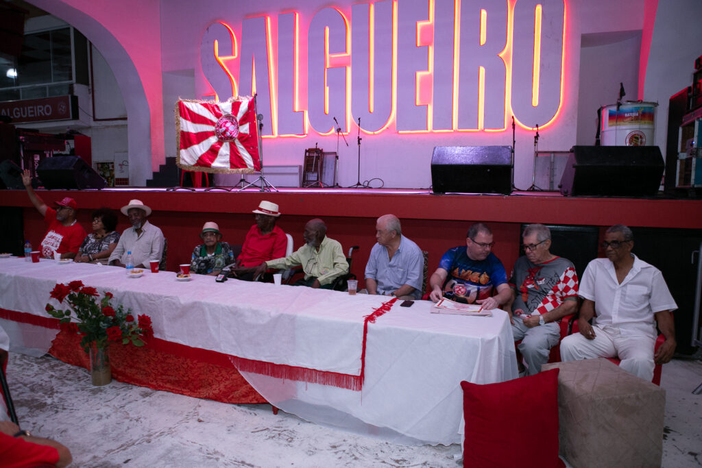 Imagem 1 - Cerimônia de posse de Djalma Sabiá como Presidente de Honra do Acadêmicos do Salgueiro. Ele está centrado no centro de uma grande mesa com uma toalha branca que está dentro da quadra do Salgueiro, em frente ao palco. Atrás aparece o nome da escola em um grande letreiro em neon vermelho. NO palco onde está o letreiro há uma bandeira da escola, alguns equipamentos de captação de audio.