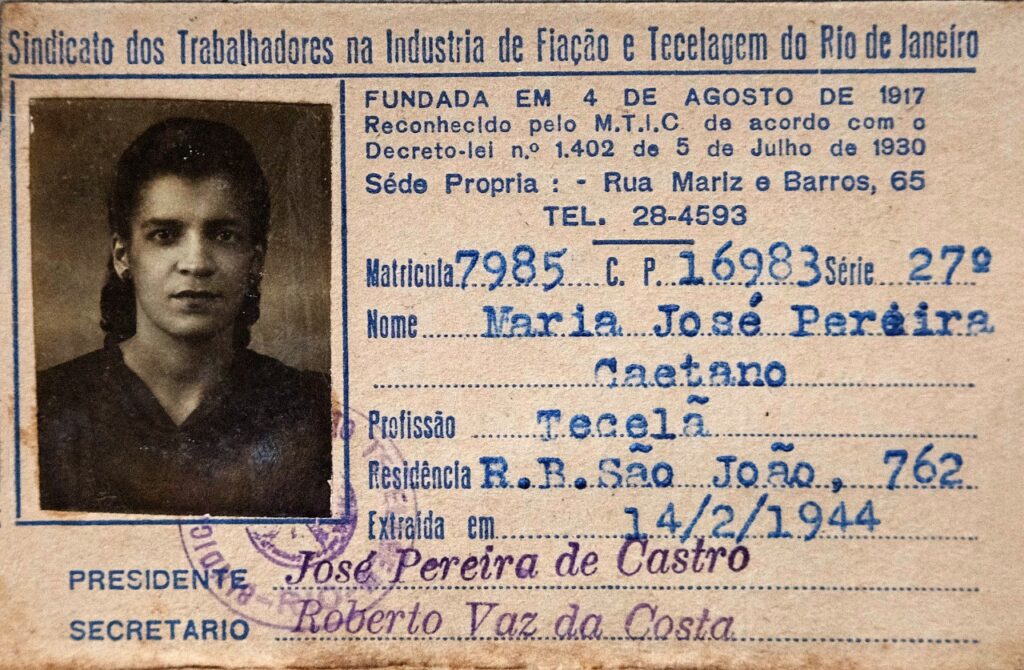 Imagem 2 - Carteira do sindicato dos trabalhadores da industria de fiação e tecelagem do Rio de Janeiro pertencente a Maria José Pereira Caetano, profissão tecelã de 14 de fevereiro de 1944. Fodo de uma mulher negra, jovem, cabelos pretos e compridos, penteados para trás e presos. Ela veste uma blusa escura, quase preta, a imagem é sépia. 